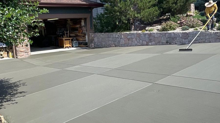 Freshly poured concrete driveway beside a house and stone wall, with trees in the background