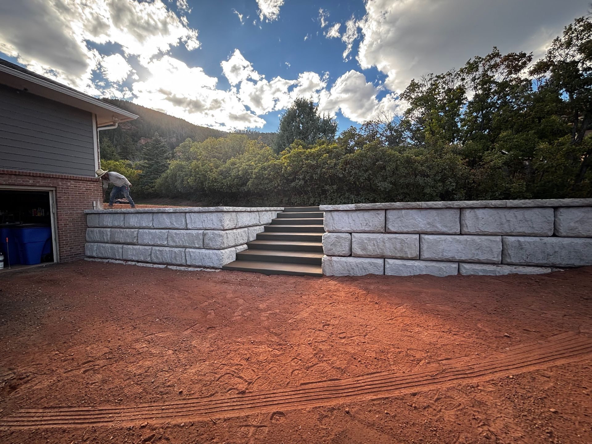 Stone steps in a red dirt courtyard beside a house, with a low retaining wall and cloudy sky