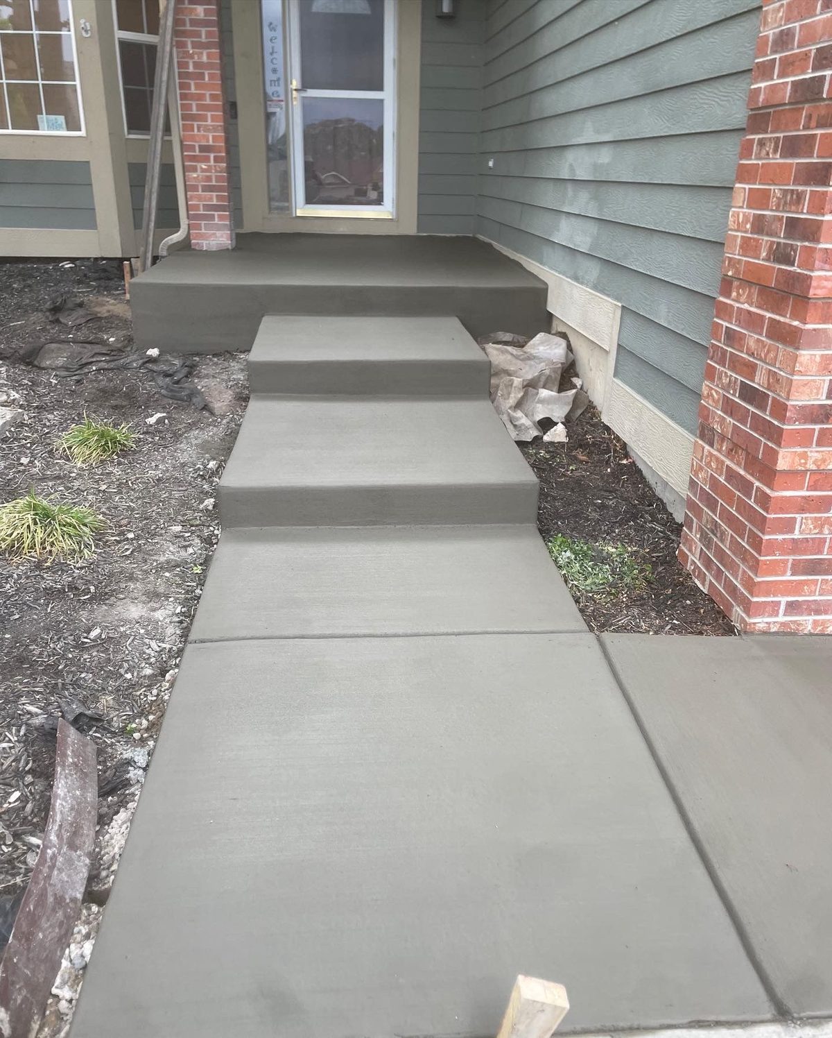 Fresh concrete walkway and three steps leading to a front porch beside brick and siding, with a small dog by the wall